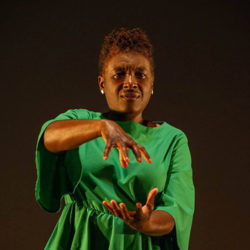 A Black woman in a bright green dress poses against a black background.