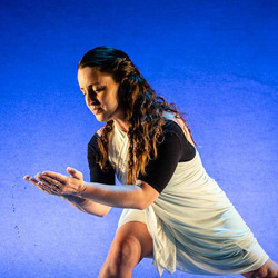 White woman in a white costume poses on stage against a blue background.