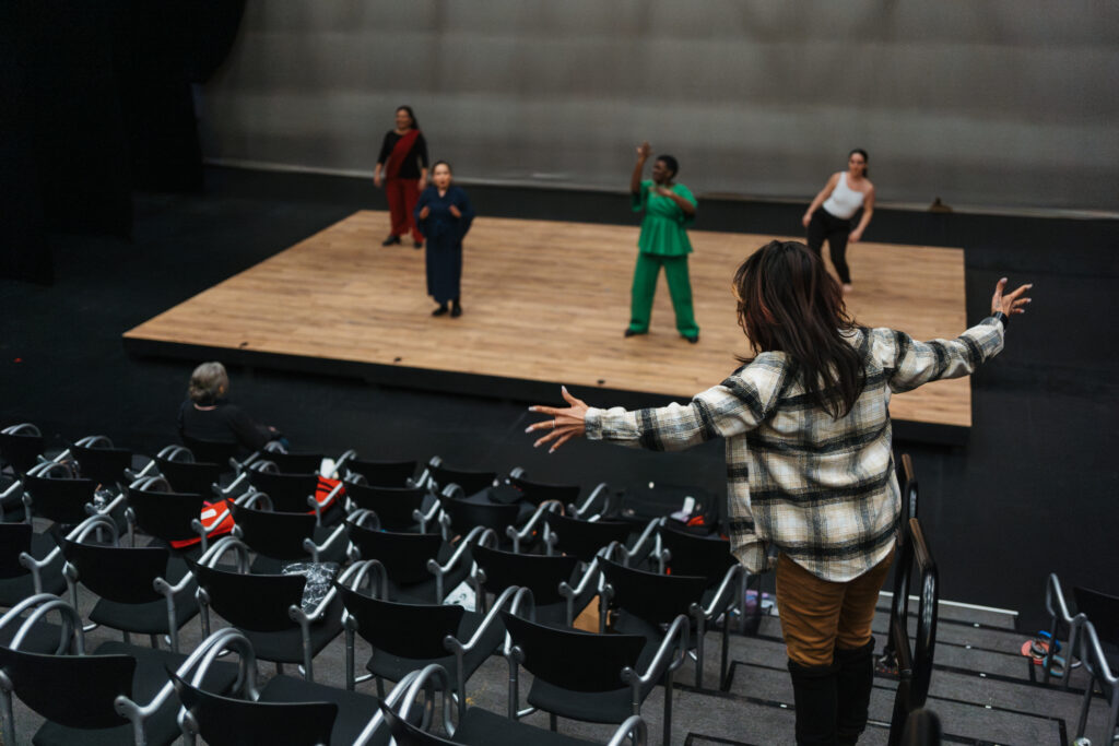 Speaking Vibrations performers in tech rehearsal. Their director is giving them instructions while they stand on stage. Photo taken from perspective of audience.