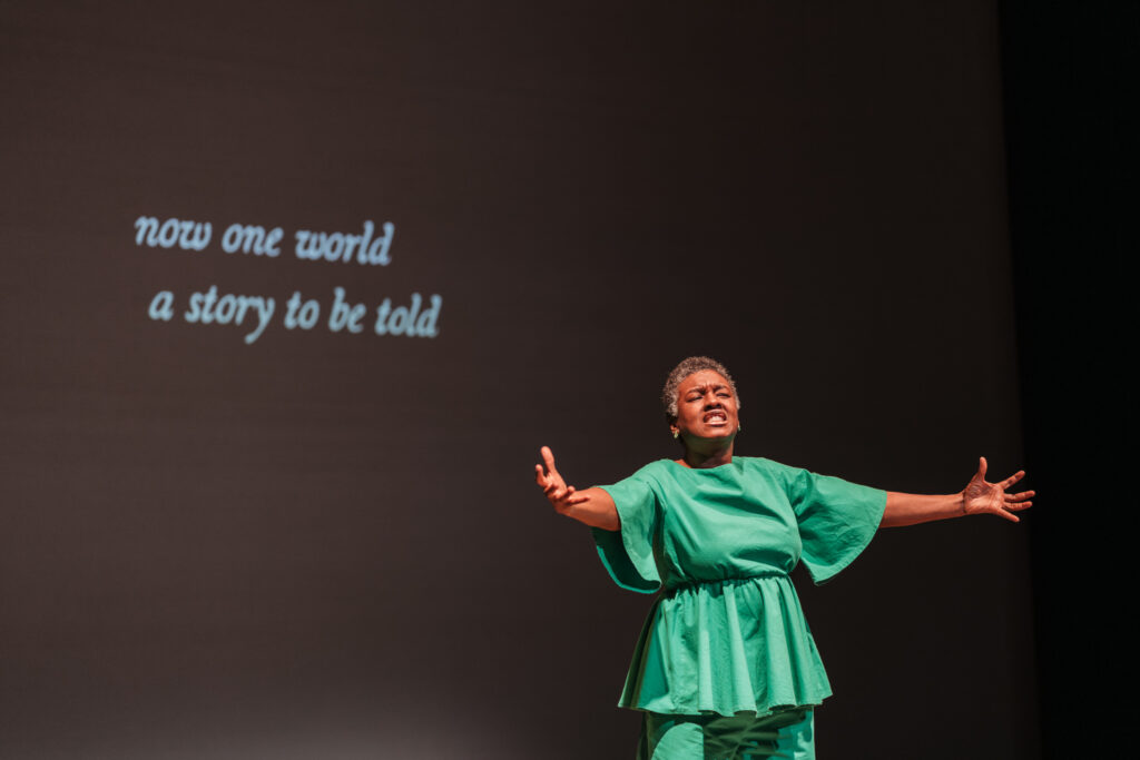 Jo-Anne Bryan, Speaking Vibrations artist on stage with her arms gestured wide open. She is in front of a caption that reads "now one world, a story to be told."